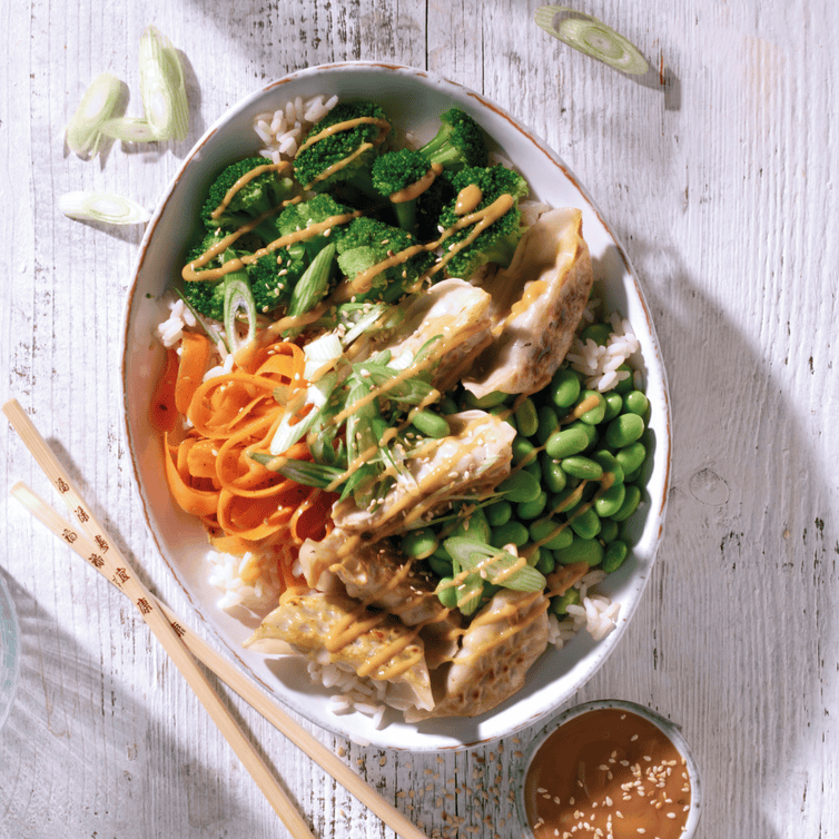 Bowl estiva con gyoza di pollo, riso, broccoli, edamame e carote con salsa alle arachidi, servita in ciotola asiatica colorata.
